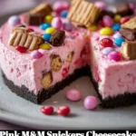 Pink M&M Snickers Cheesecake topped with colorful candies and chocolate drizzle