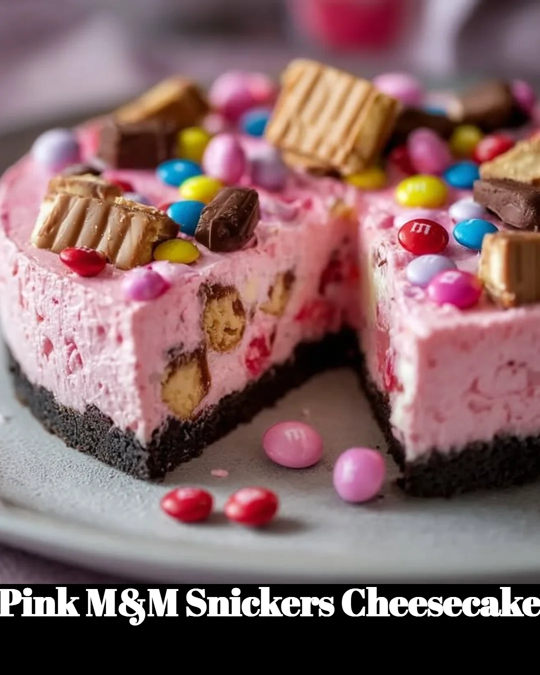 Pink M&M Snickers Cheesecake topped with colorful candies and chocolate drizzle